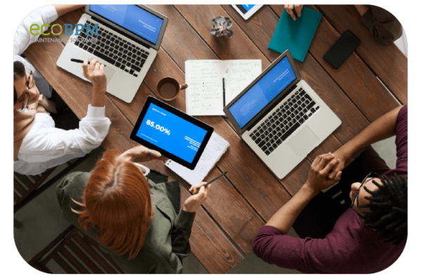 Overhead view of a business meeting with people using laptops and a tablet displaying data around a wooden table.